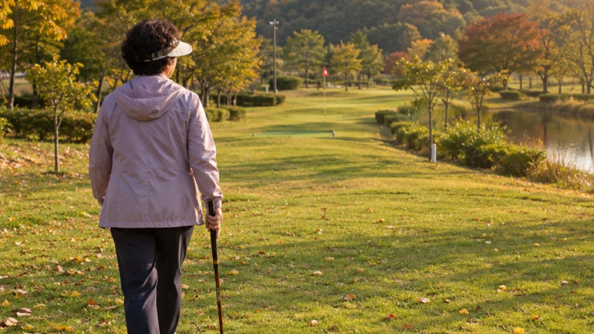 단풍이 든 언덕 아래 가을 파크골프장에서 클럽 하나를 들고 다음 홀로 걸어가는 60대 여성의 뒷모습 (AI 장면)